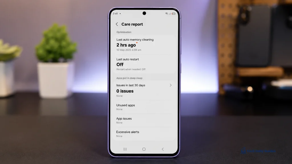 Lavender Galaxy A37 5G standing upright on a dark professional tech table with the Care report screen open, showing memory cleaning, auto restart, unused apps, app issues, and excessive alerts, with a blurred plant and modern tech workspace in the background.